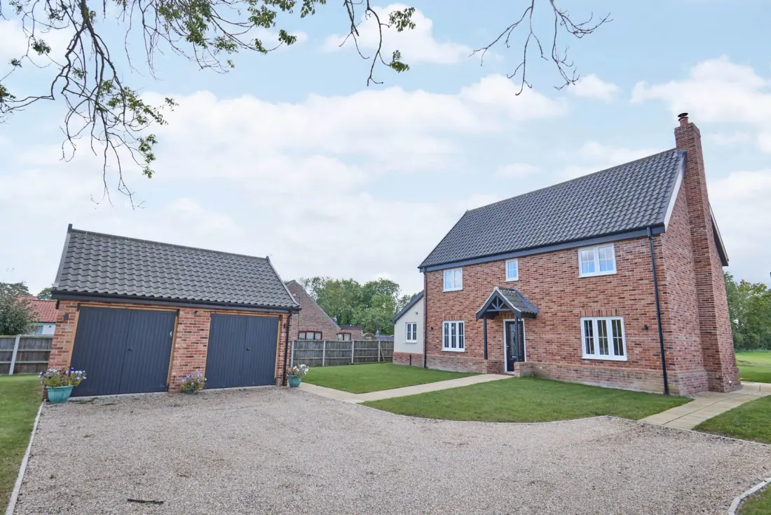 The home from the front with the gravel driveway and detached double garage in view, mature trees framing the approach