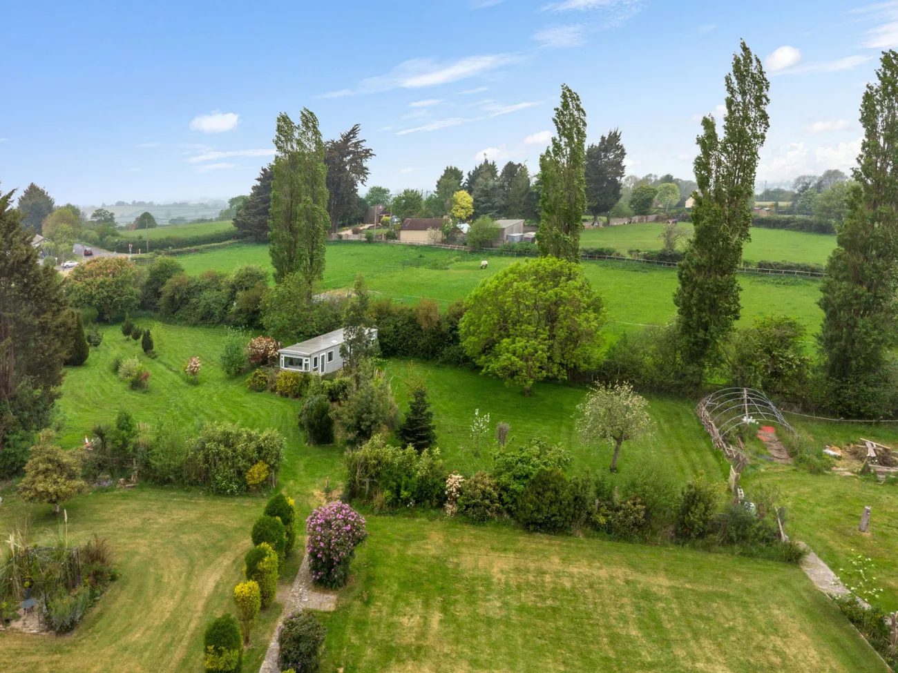 Aerial of the lower garden showing The Retreat lodge, fruit trees, and the polytunnel framework, with paddocks and far fields beyond.